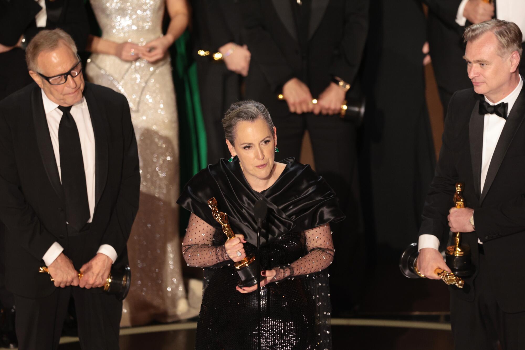 A woman flanked by two men holding an Oscar statuette on stage.