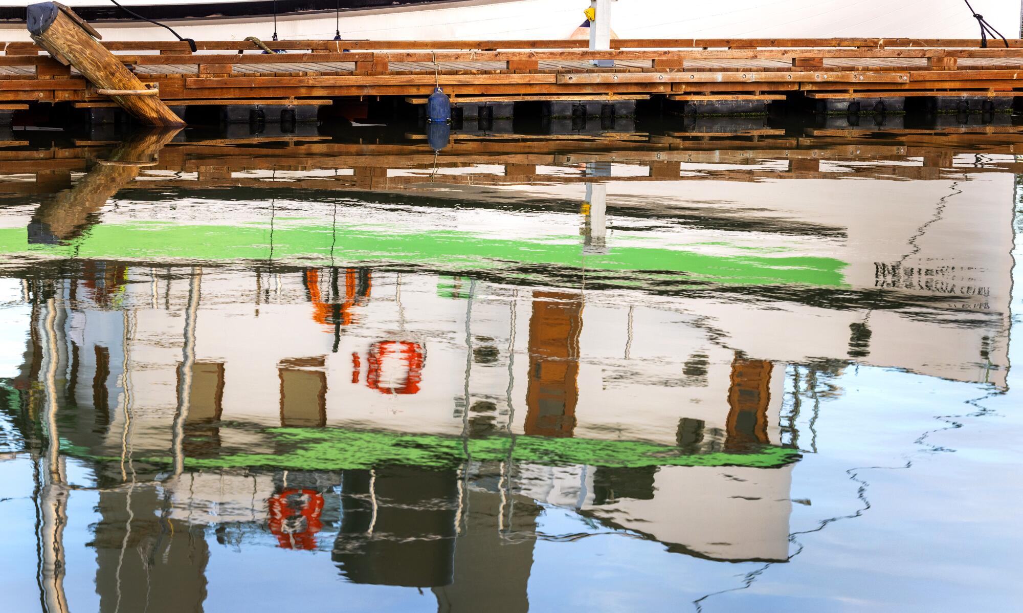 A reflected image of a green and white boat in the water alongside a pier.