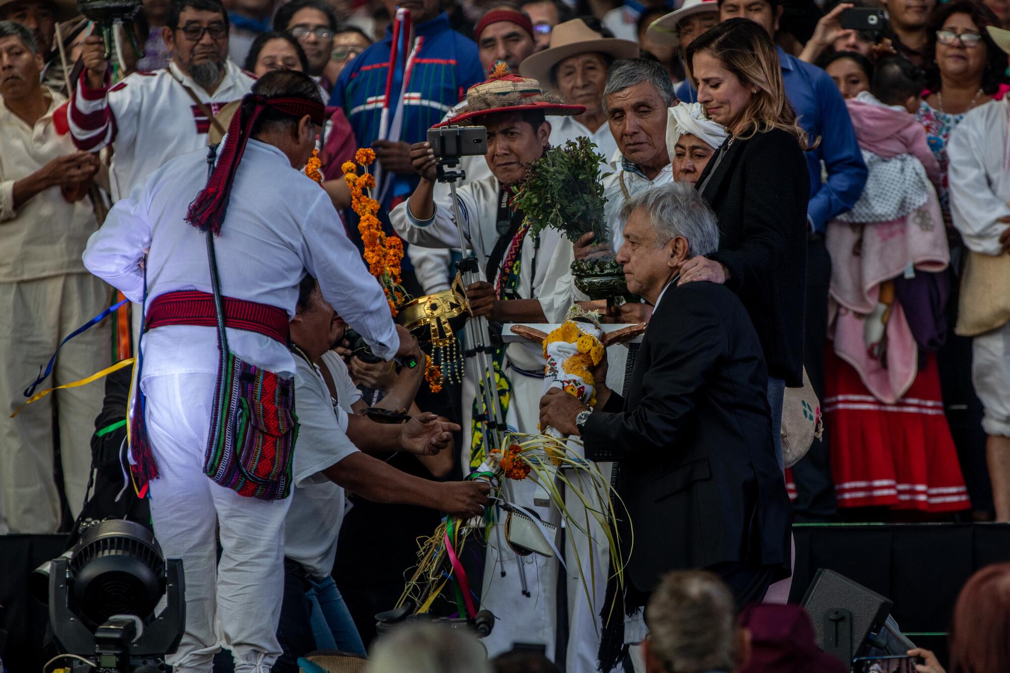 A man in a suit kneels facing a group of people dressed in white, some holding plants and flowers