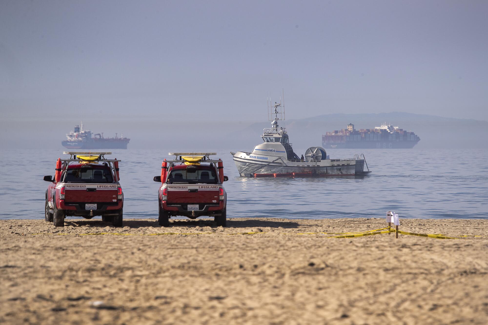 Lifeguard vehicles face the water at Huntington Beach while oil-containing booms are put in place by boats offshore.