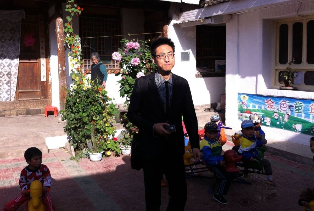 A bespectacled man in suit and tie walks in a courtyard where children play near flowering plants 
