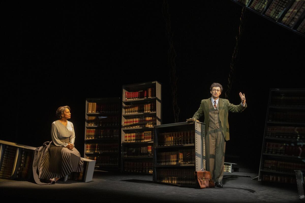 A seated woman looks at a man talking with his hand raised, on a stage set with bookcases.