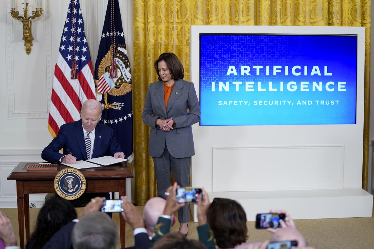 President Biden sits and signs a document as Kamala Harris stands next to him, and people take cellphone video.