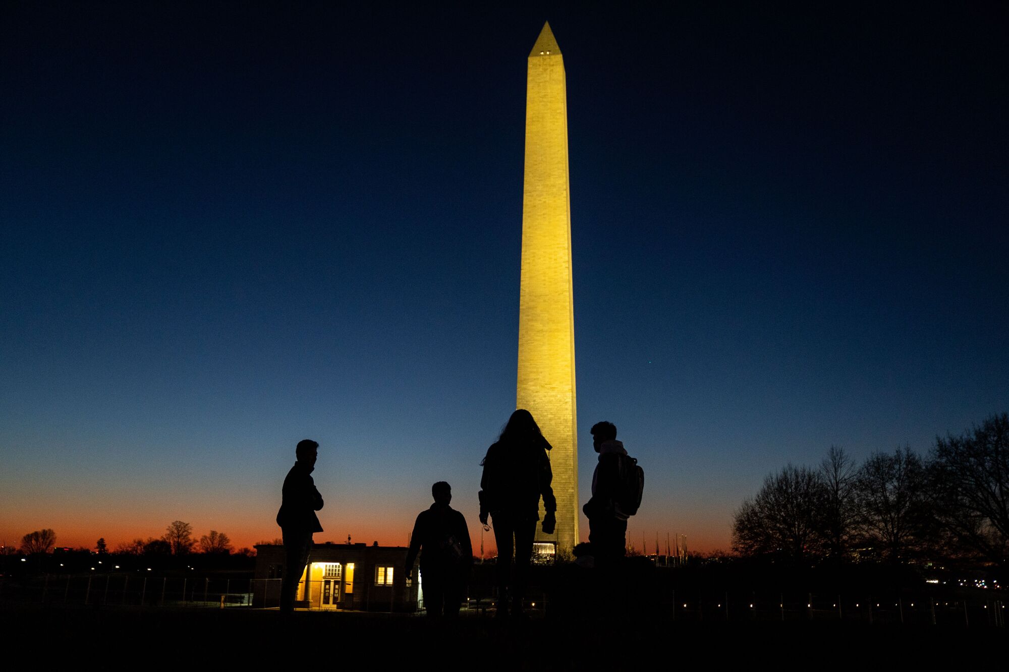People walk on the National Mall with the Washington Monument illuminated in the evening sky.