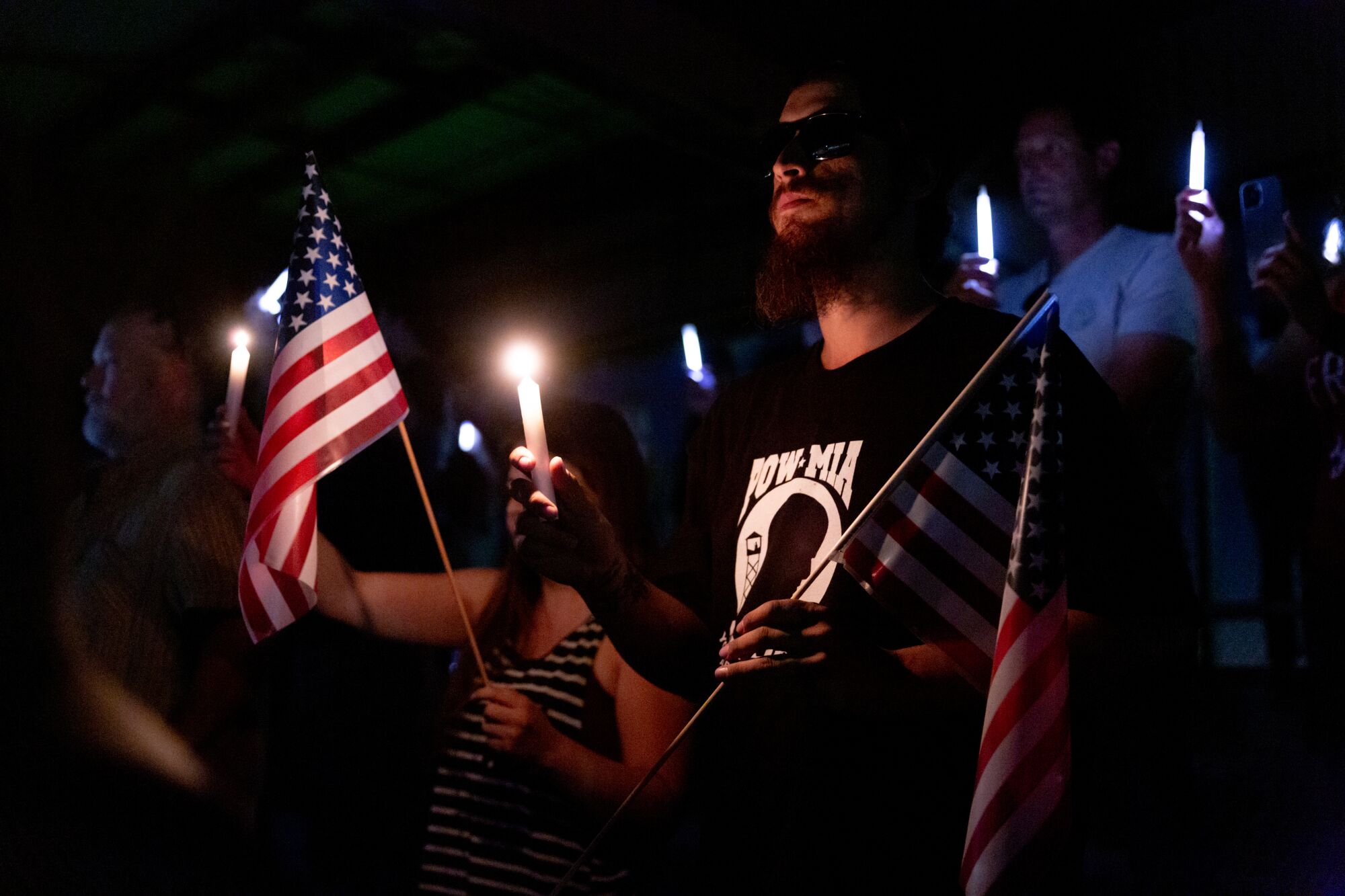 A man in a POW-MIA T-shirt holds a flag and a candle in the dark among other people.