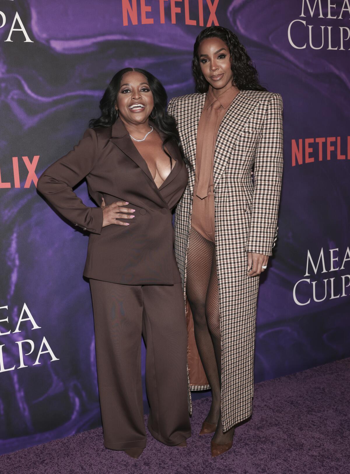 Sherri Shepherd in a brown suit standing next to Kelly Rowland in a long brown, checkered coat against a purple backdrop