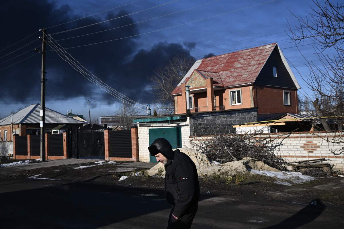 A man walks in a street as black smoke rises in the distance behind homes.
