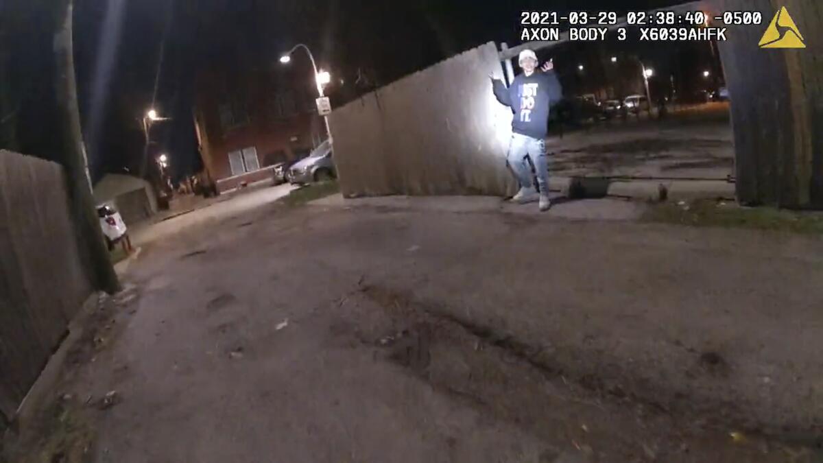 A teenage boy stands with both hands raised next to a wooden fence in an alley with a flashlight shined on him at night