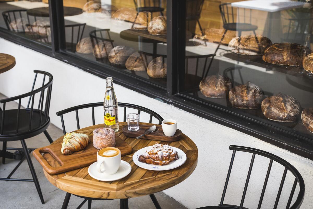 A photo of a spread of pastries atop a patio table. In the window beyond, rows of bread loaves sit on shelves.