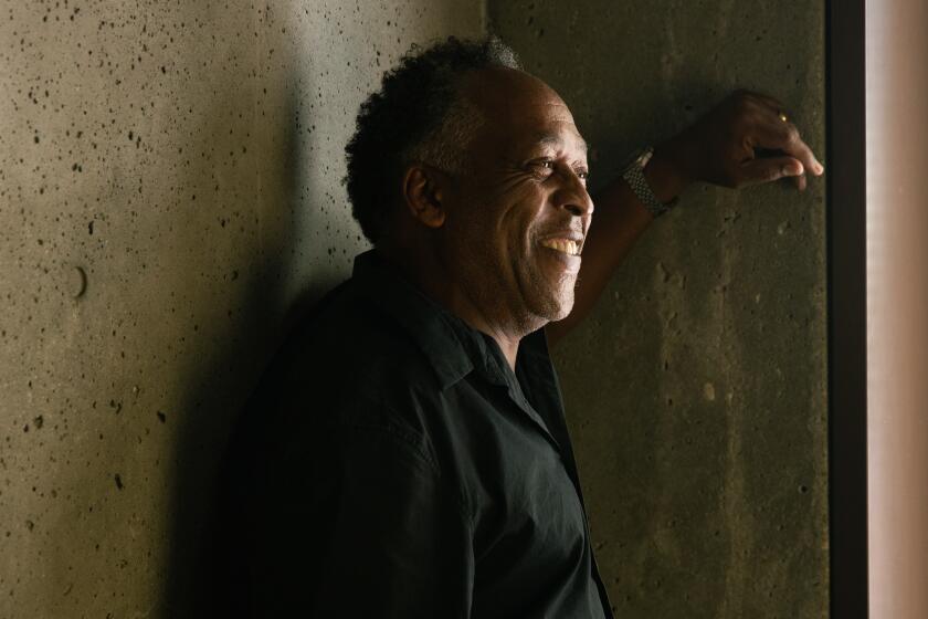 Henry Taylor, wearing a black shirt and leaning on a concrete wall, is illuminate by a small beam of sunlight 