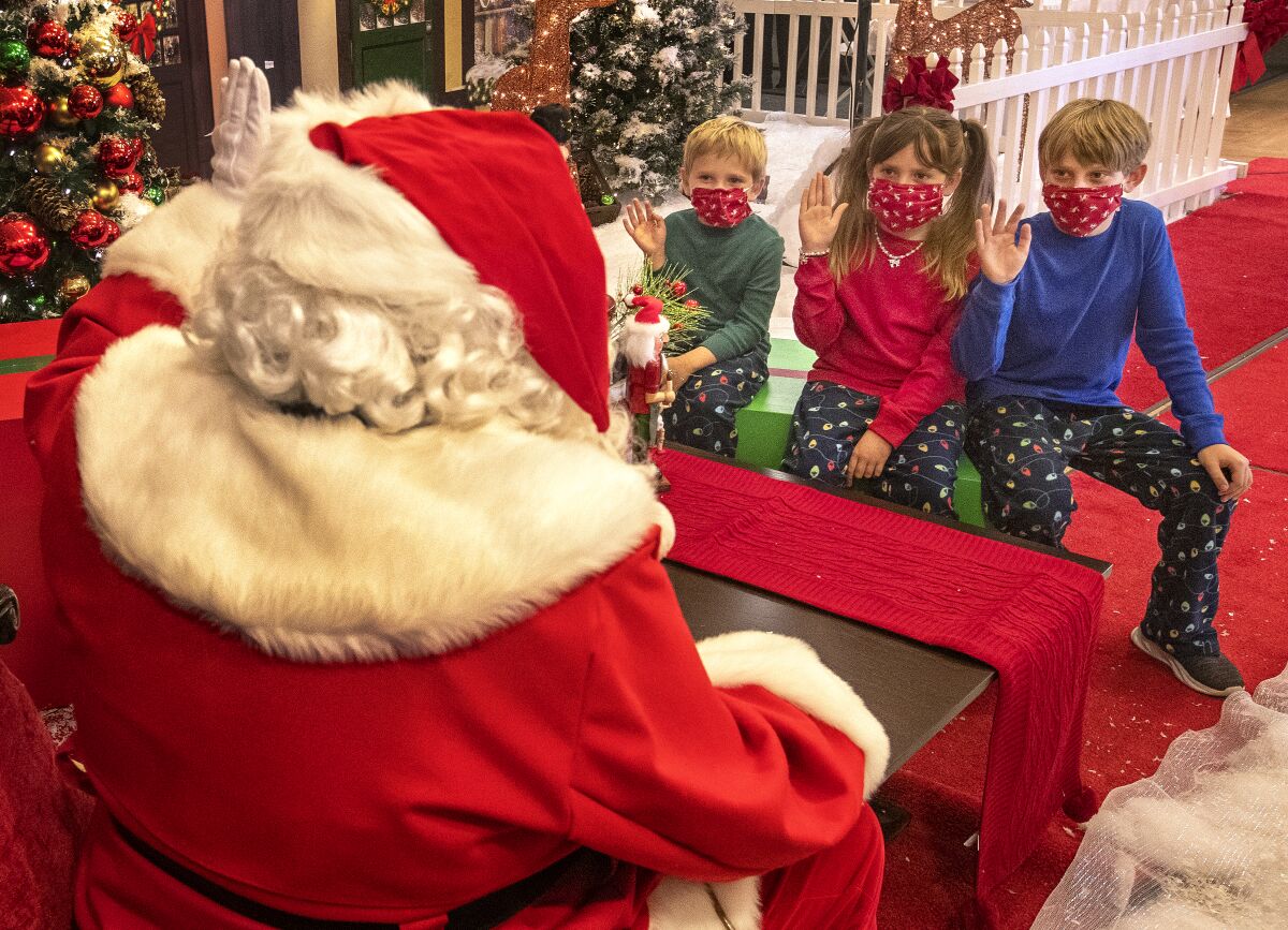 Masked kids in Christmas-themed clothes and masks sit on a bench and wave to Santa.