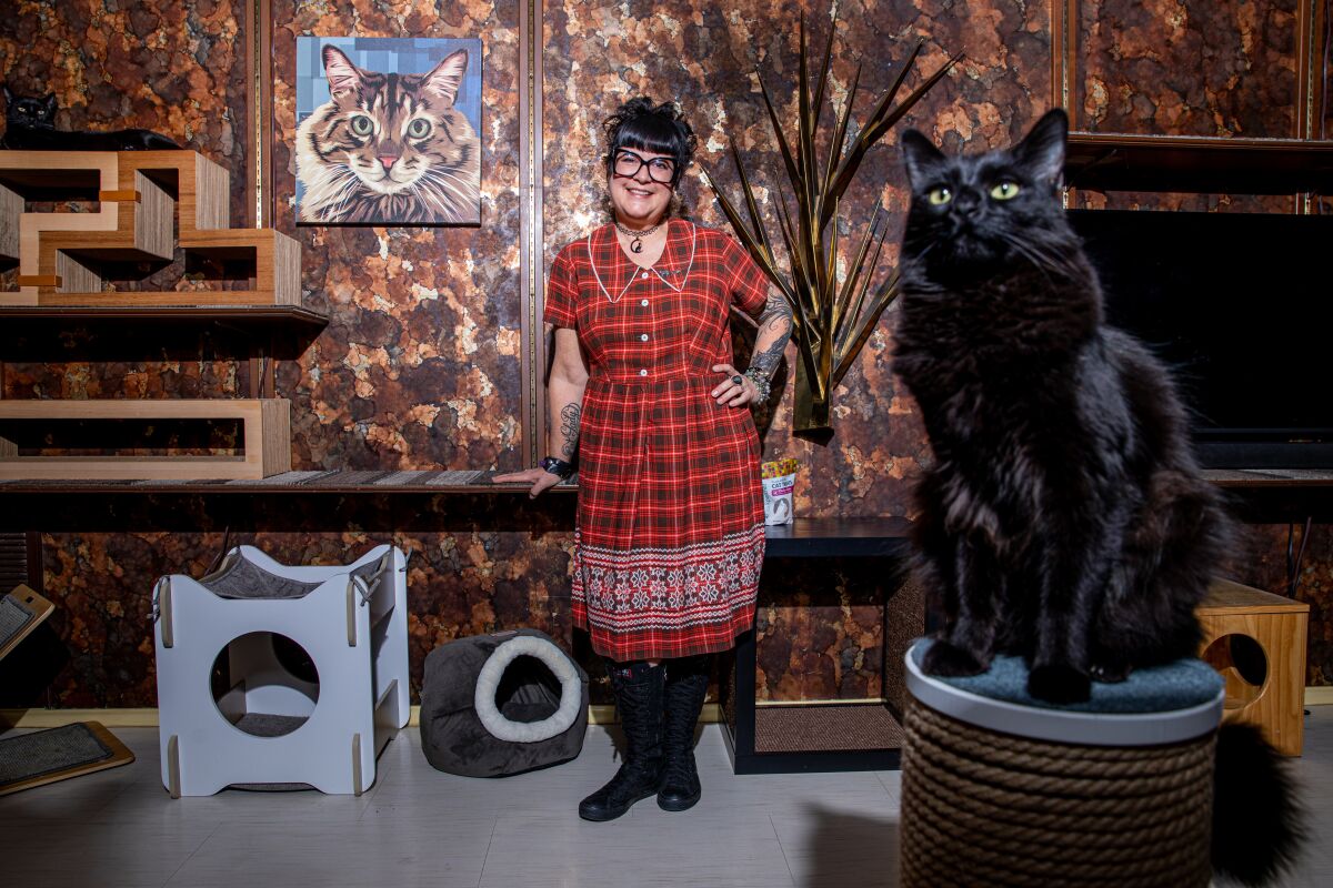 A  woman with one of her black cats, who's sitting atop a scratching post in her condo.