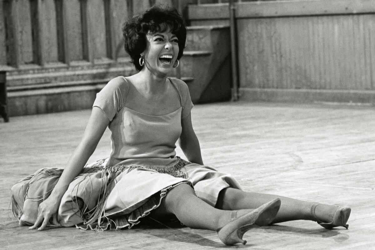 A young Rita Moreno sits laughing on a hardwood floor.