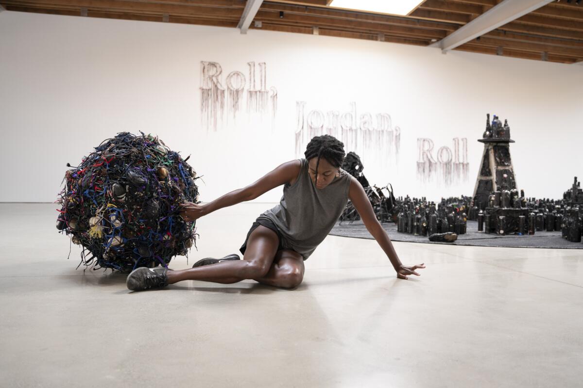 Jessican Emmanuel, in gray tank and black running shorts, drags a spherical black sculpture through a gallery space.