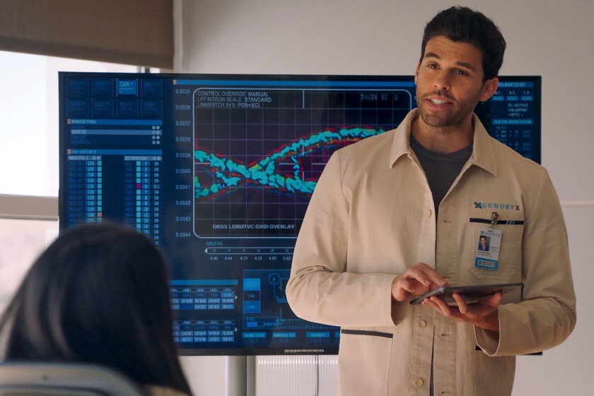 On "Roswell, New Mexico" a man wearing a security badge stands in front of a big video screen displaying a graph.