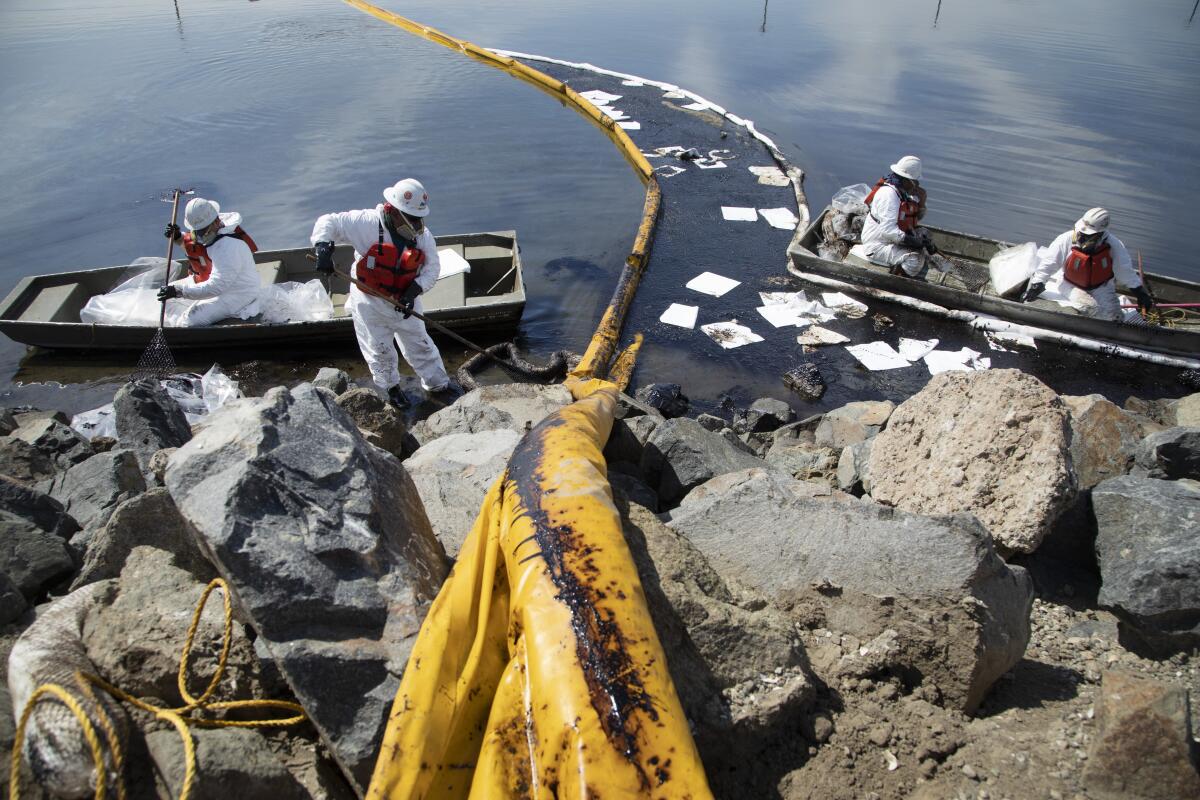 Four workers in small boats wear white suits and hold tools on a rocky coastline.