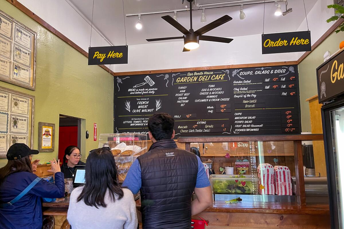 People line up at a deli, where the menu is on the wall in front of them.