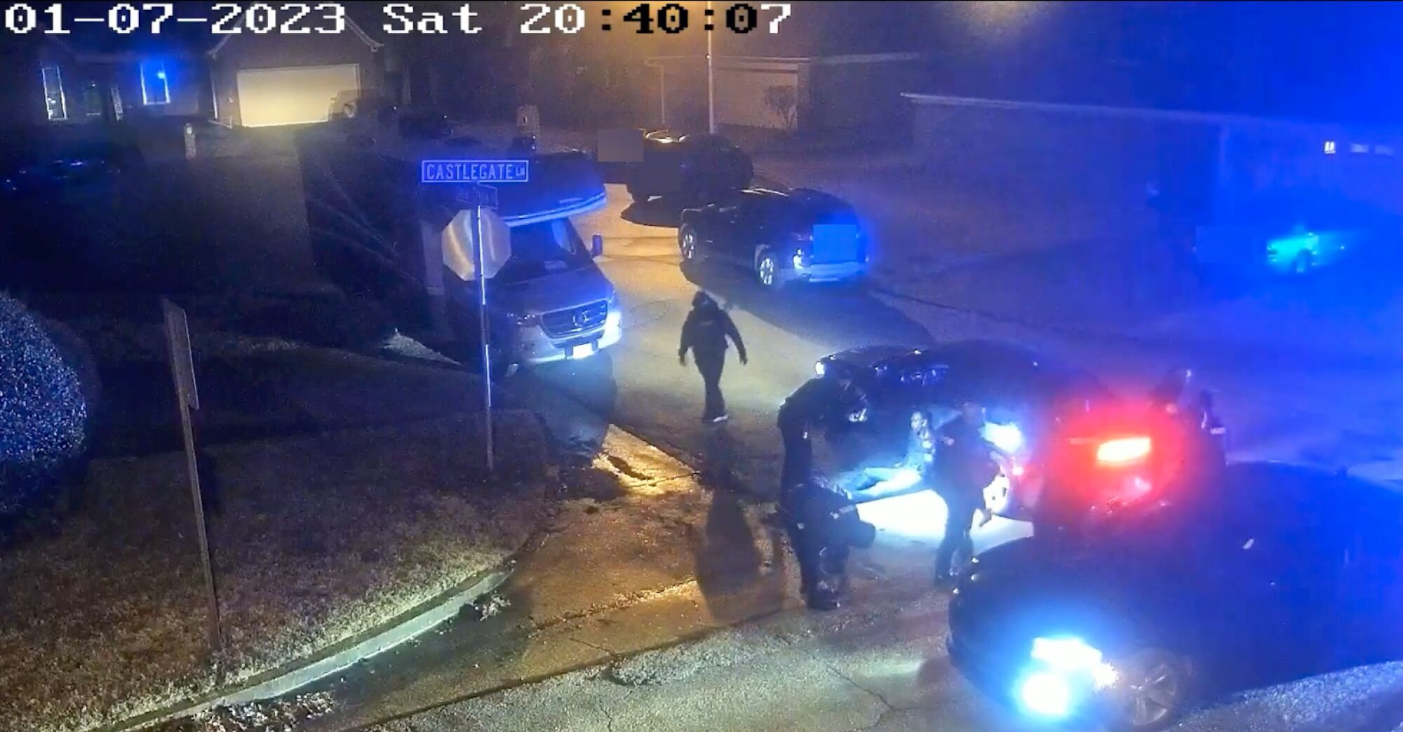 Police officers restrain a person near parked vehicles on a residential street