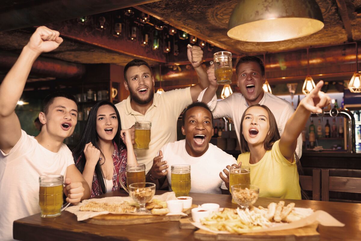 Happy friends having fun in pub watching sport in TV together, drinking beer, cheering for team. Group of people celebrating victory of favorite team, emotionally gesturing hands and shouting goal.