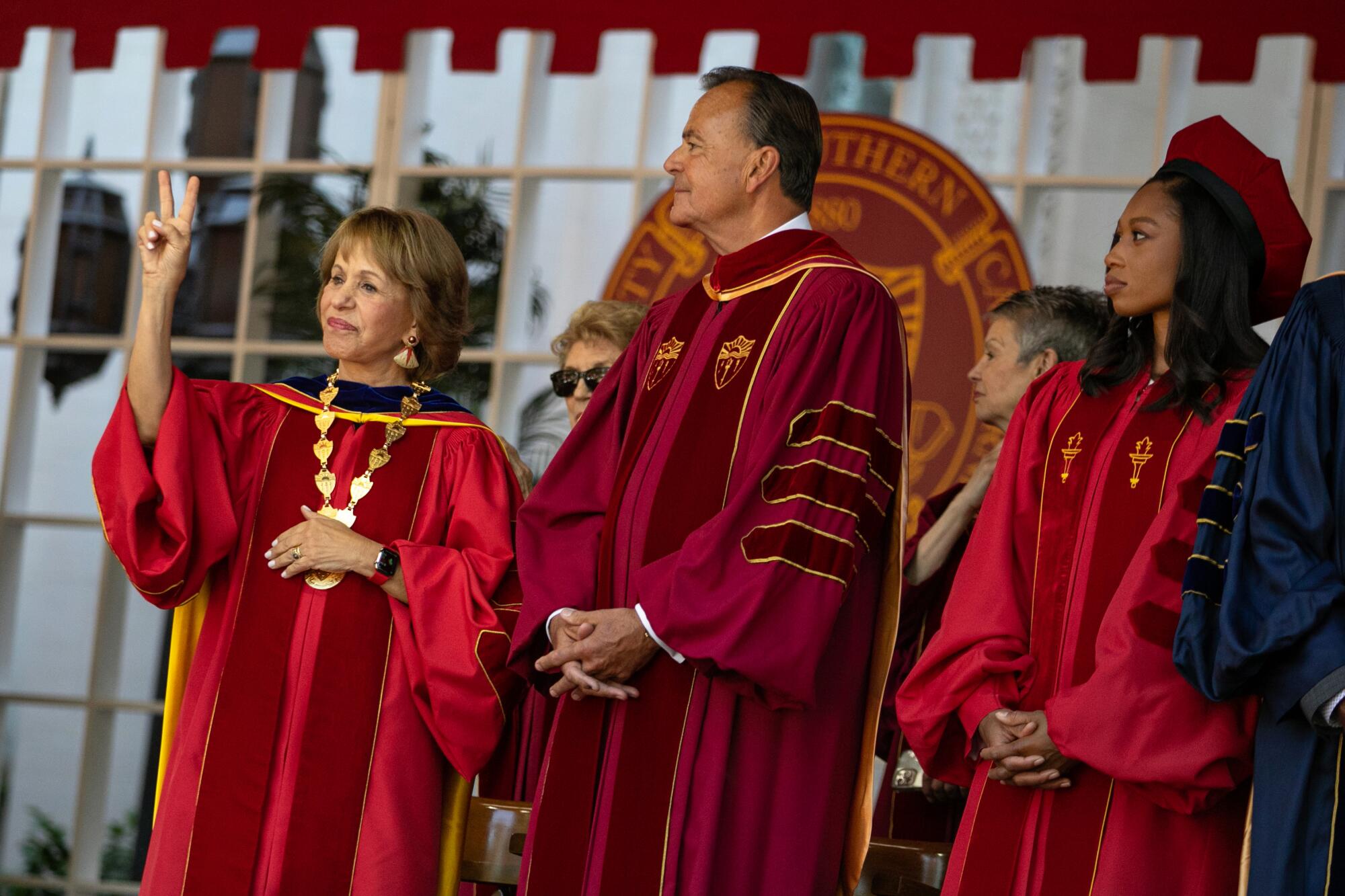 People in robes stand on stage, including a woman who holds two fingers up