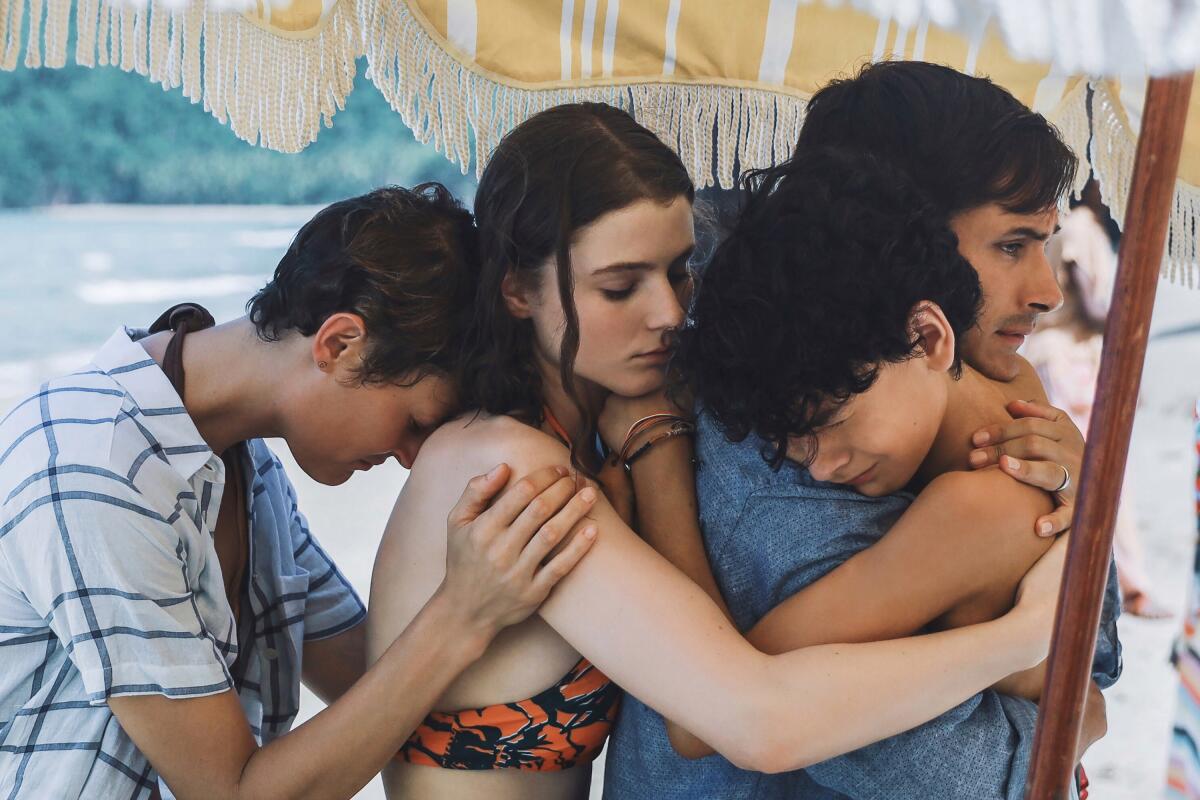 A group of emotional people embrace beneath a beach umbrella.