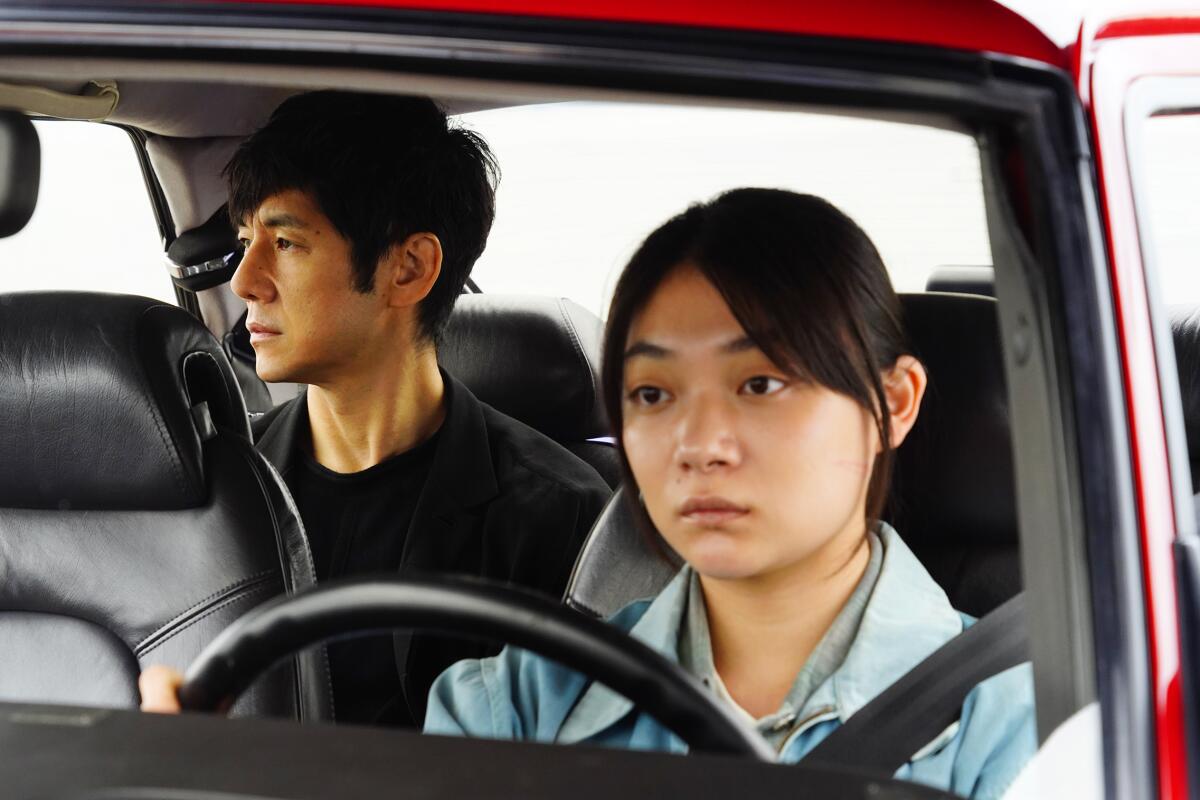 A woman sits behind the wheel of a car with a man in the back