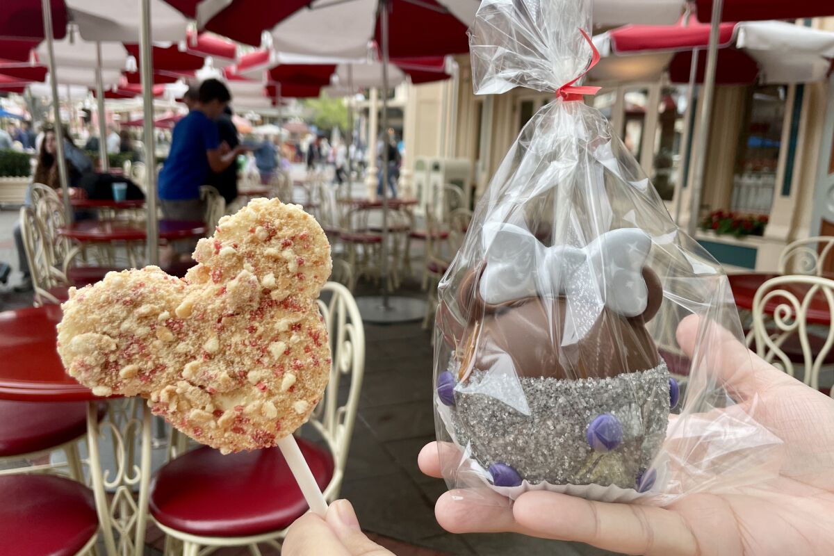 Two hands hold a strawberry-shortcake marshmallow and a chocolate and caramel apple from Disneyland.