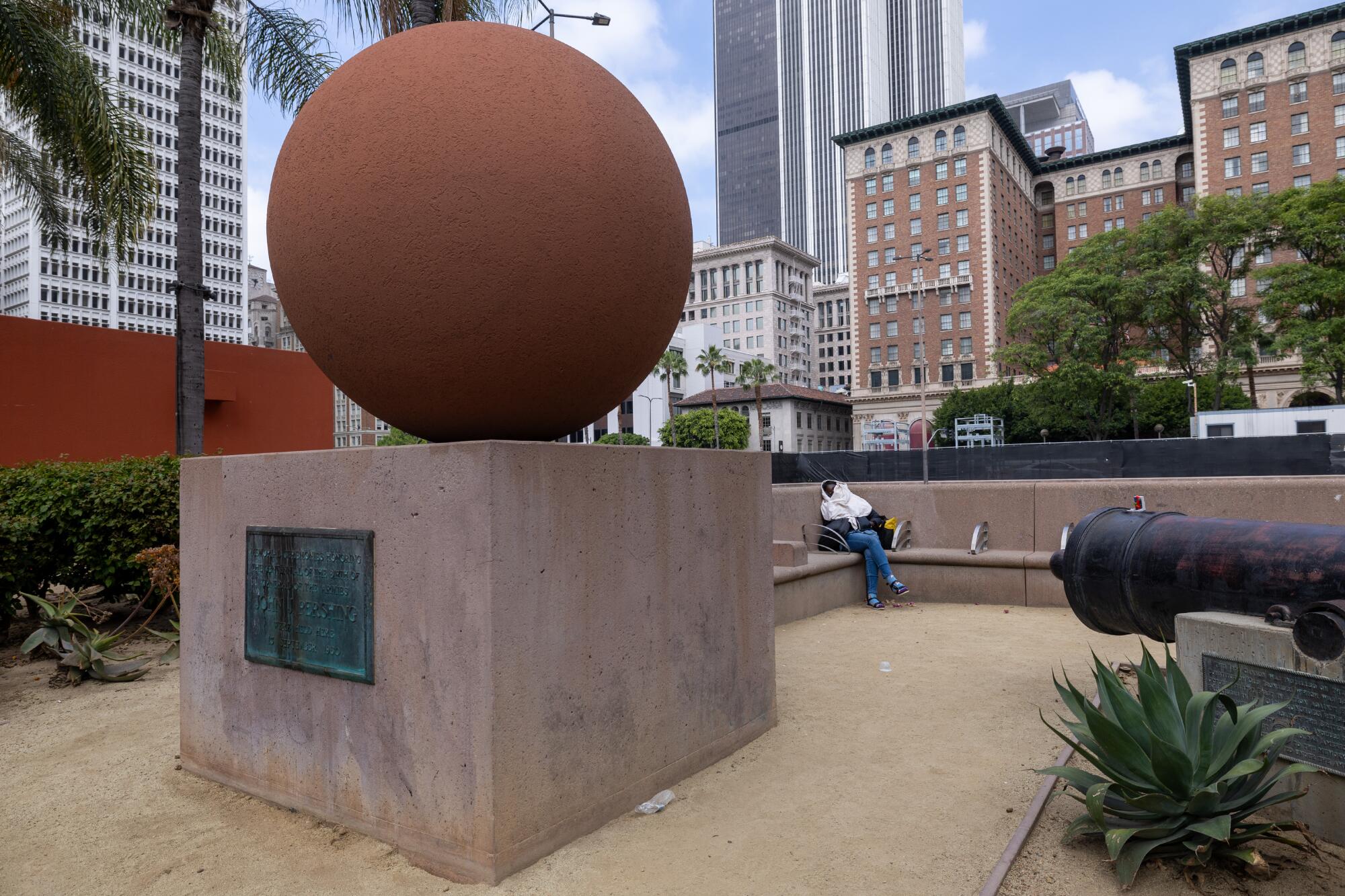 A woman napping on a bench behind a sculpture of a sphere on a square base, with high-rises as a backdrop