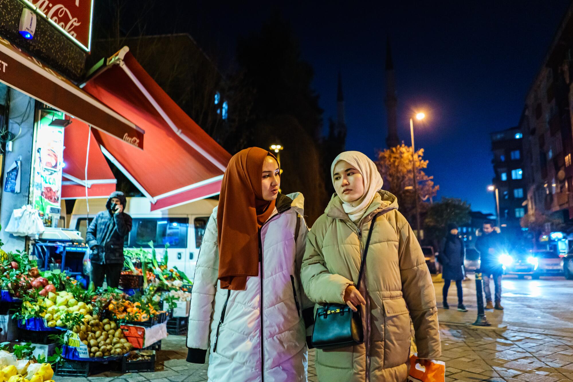 Two young women in head coverings and puffer coats walk past a produce stand on a street 