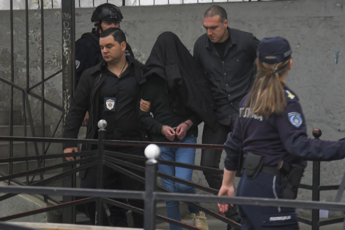 A person in handcuffs, with head covered, is escorted by people in dark clothing  