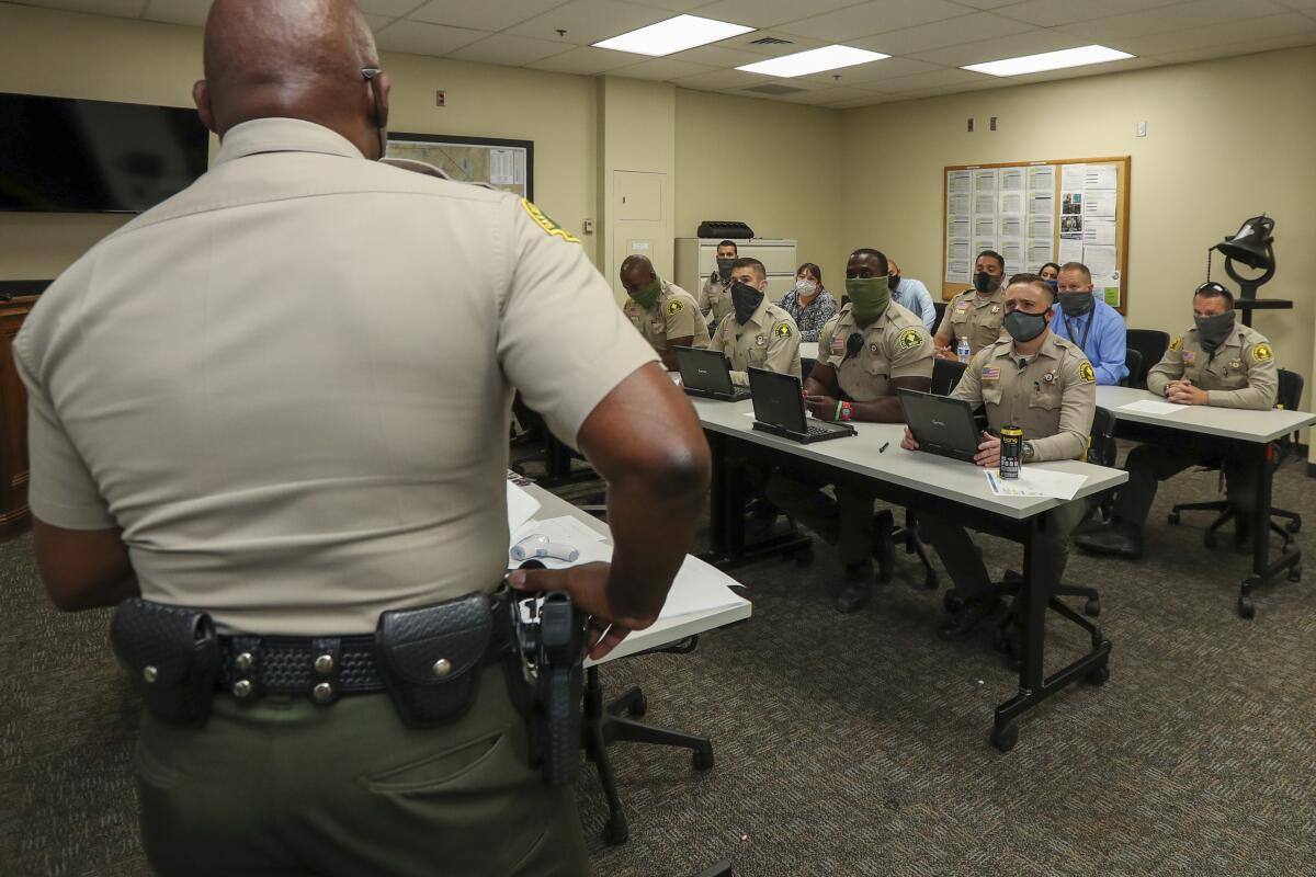 A group of men wearing tan shirts and masks sitting in rows of tables with laptops.