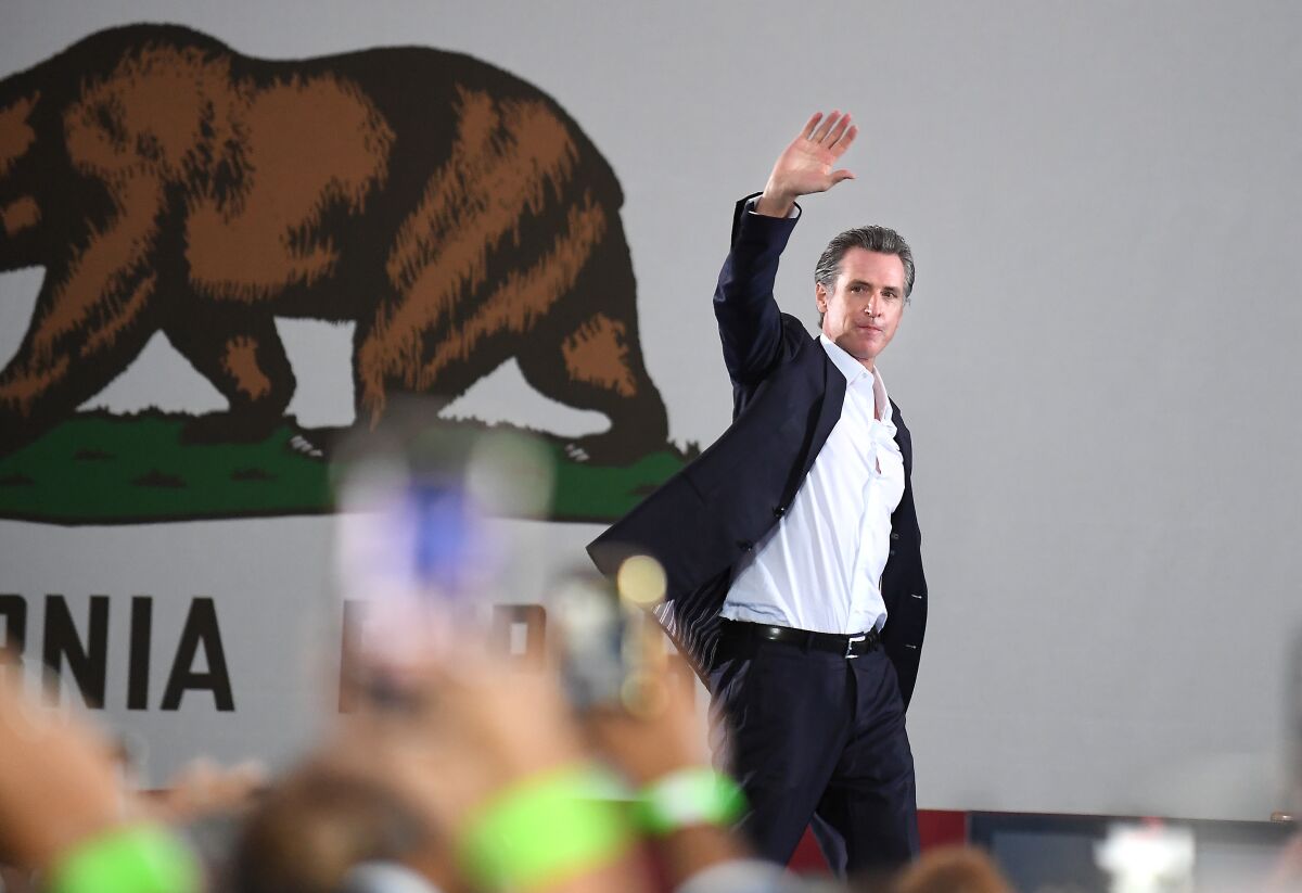 Gov. Gavin Newsom, standing before a large California state flag, waves from a stage.