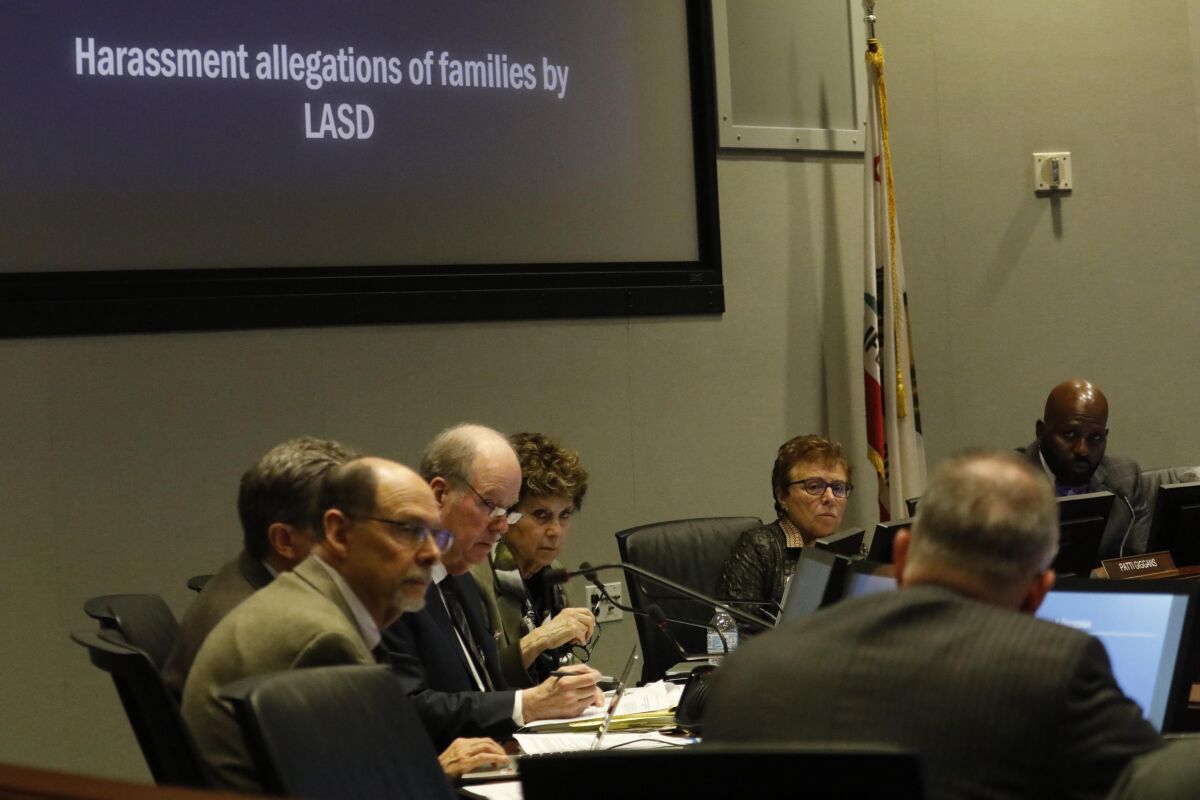 People sit at a conference table under a screen reading "Harassment allegations of families by LASD."