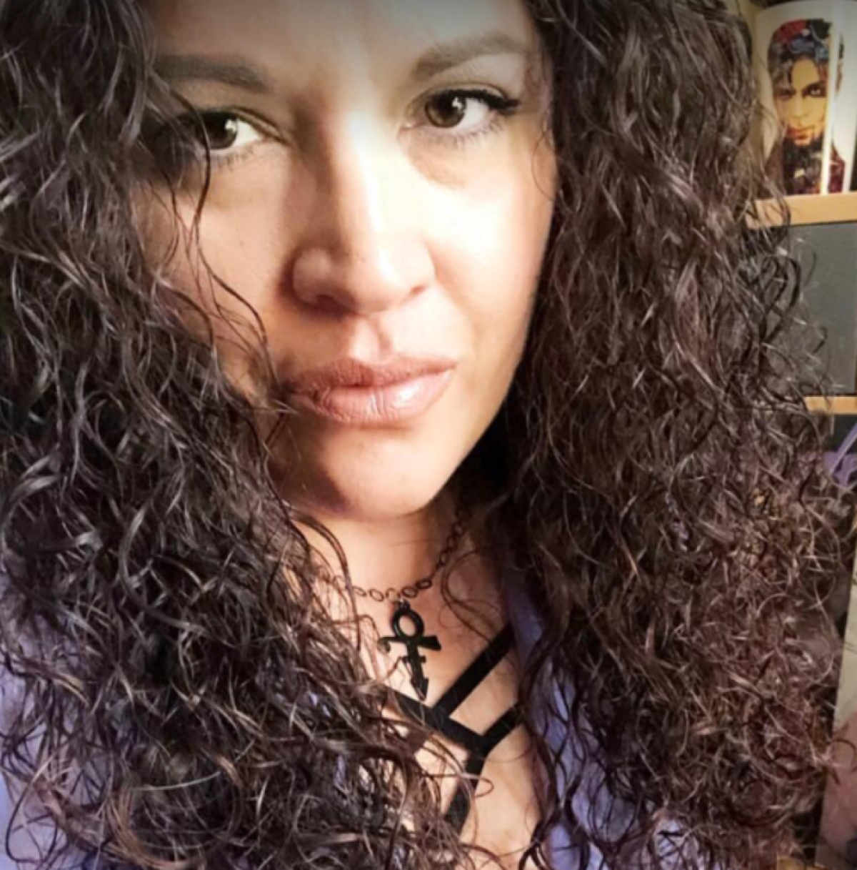 Woman with curly brown hair wears purple shirt and black Prince Love Symbol necklace. 