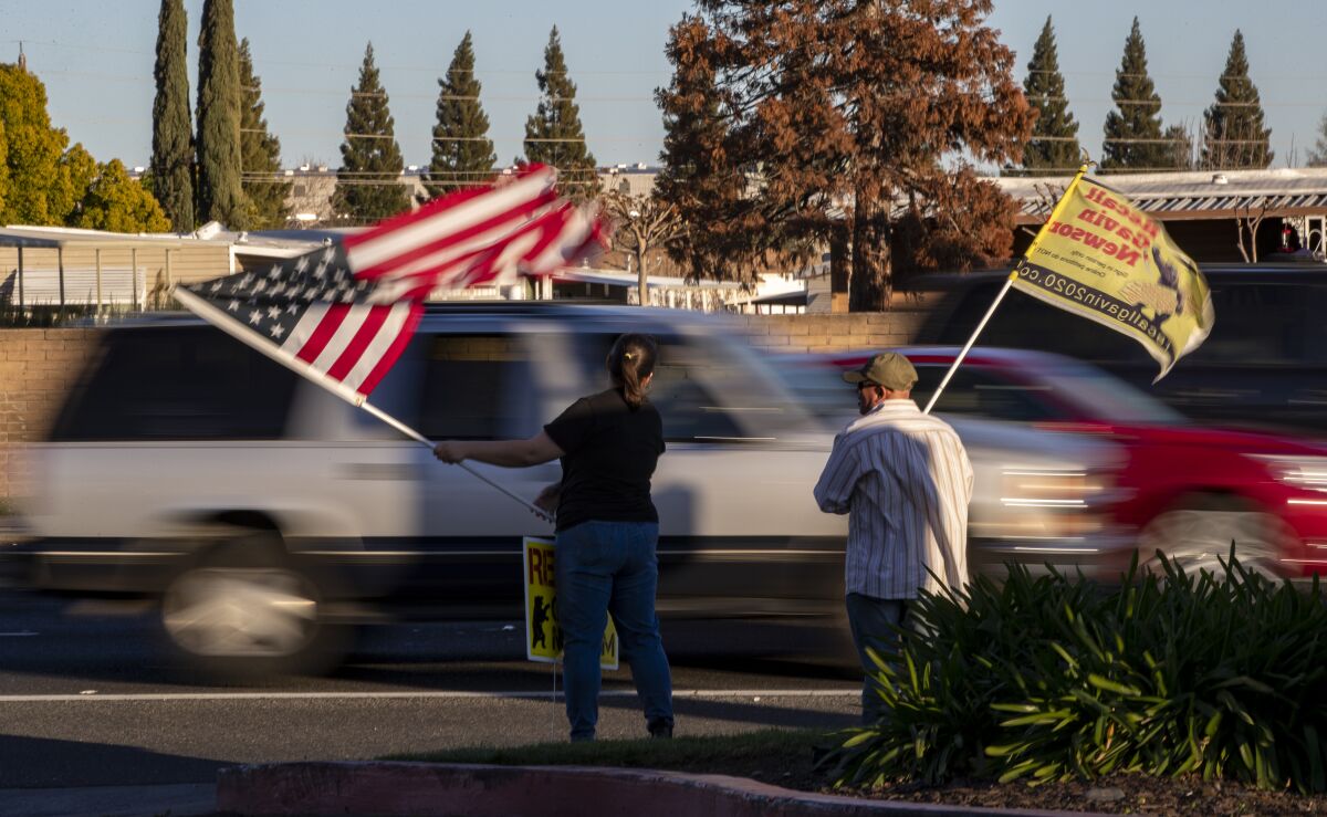 Two people wave an American flag and a "Recall Gavin Newsom" flag on the side a road.