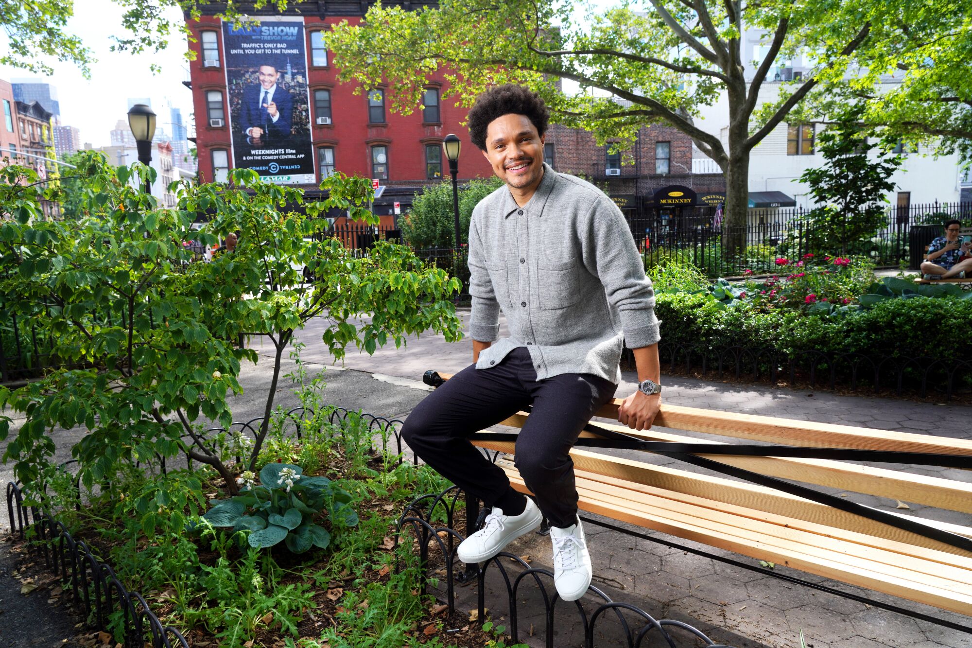 A smiling Trevor Noah sits on the back of a park bench in New York City.