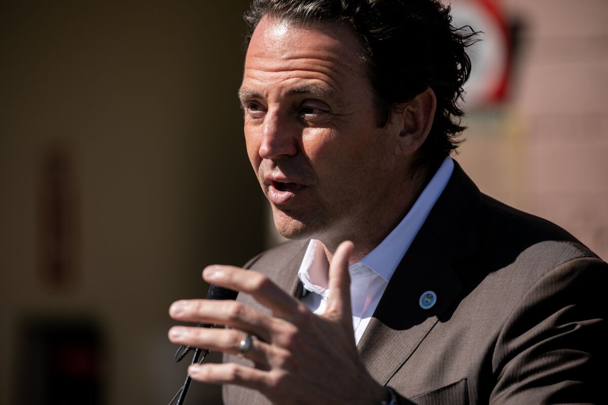 A man in a suit jacket speaks, his face lit by sharp sunlight, and gestures.