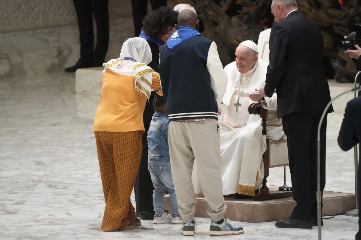 Pope Francis sits while people stand in front of him in a hall.