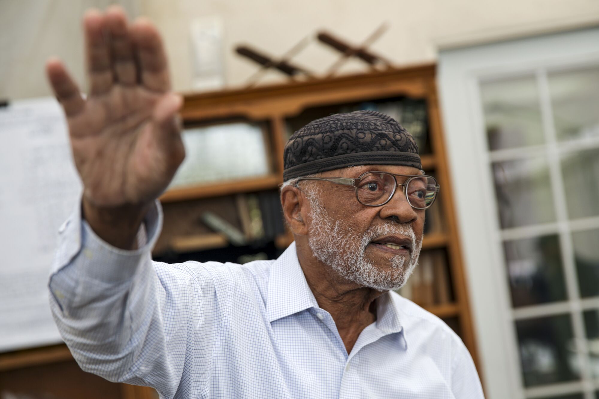 An older man wearing a cap and glasses with his hand raised