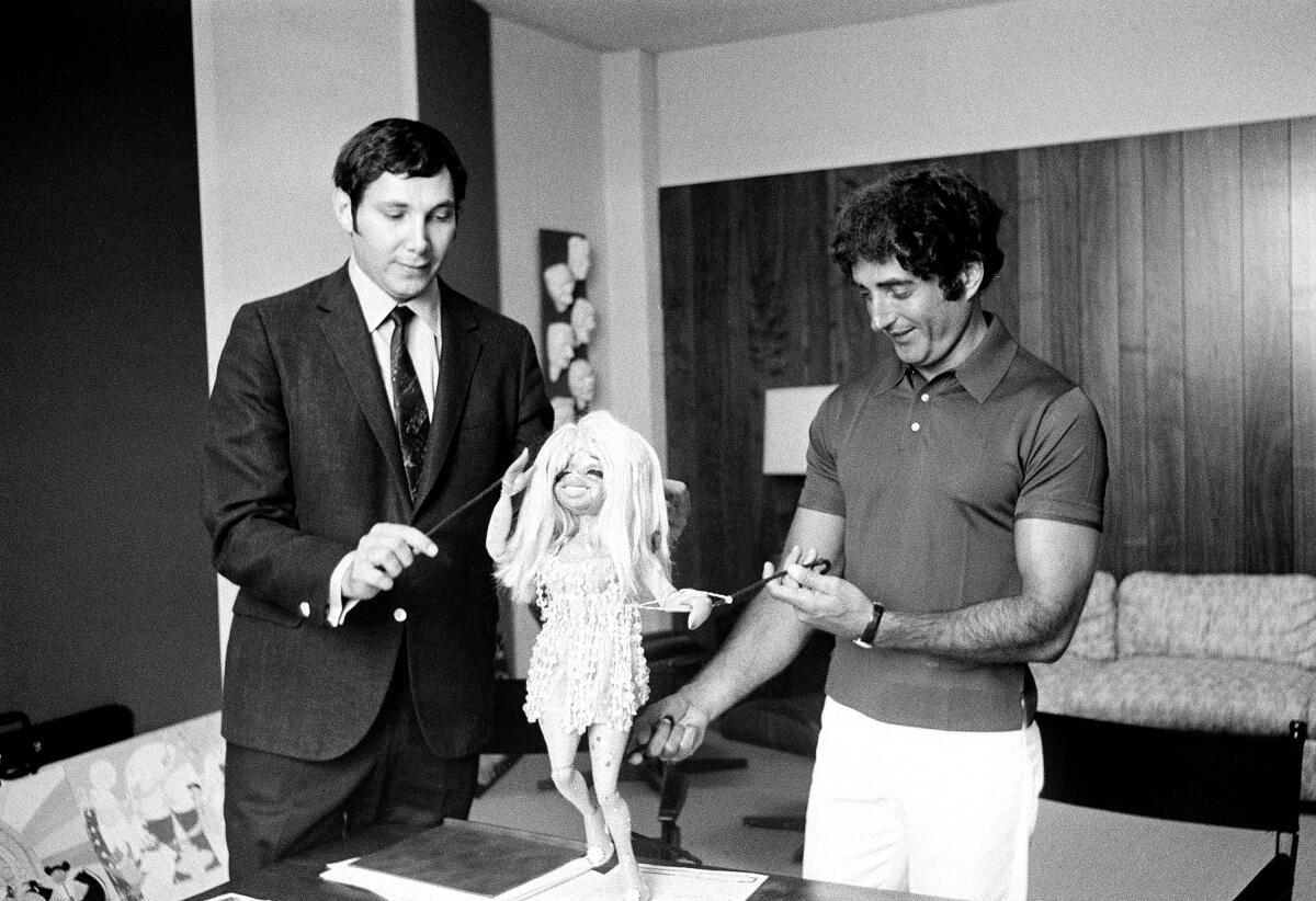 Marty Krofft and Sid Krofft stand on either side of a puppet standing on a table.