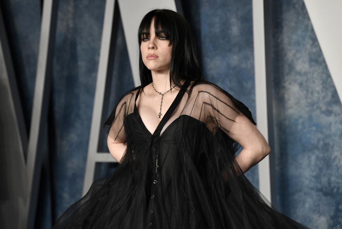 Billie Eilish with black hair posing in a black gown with sheer sleeves