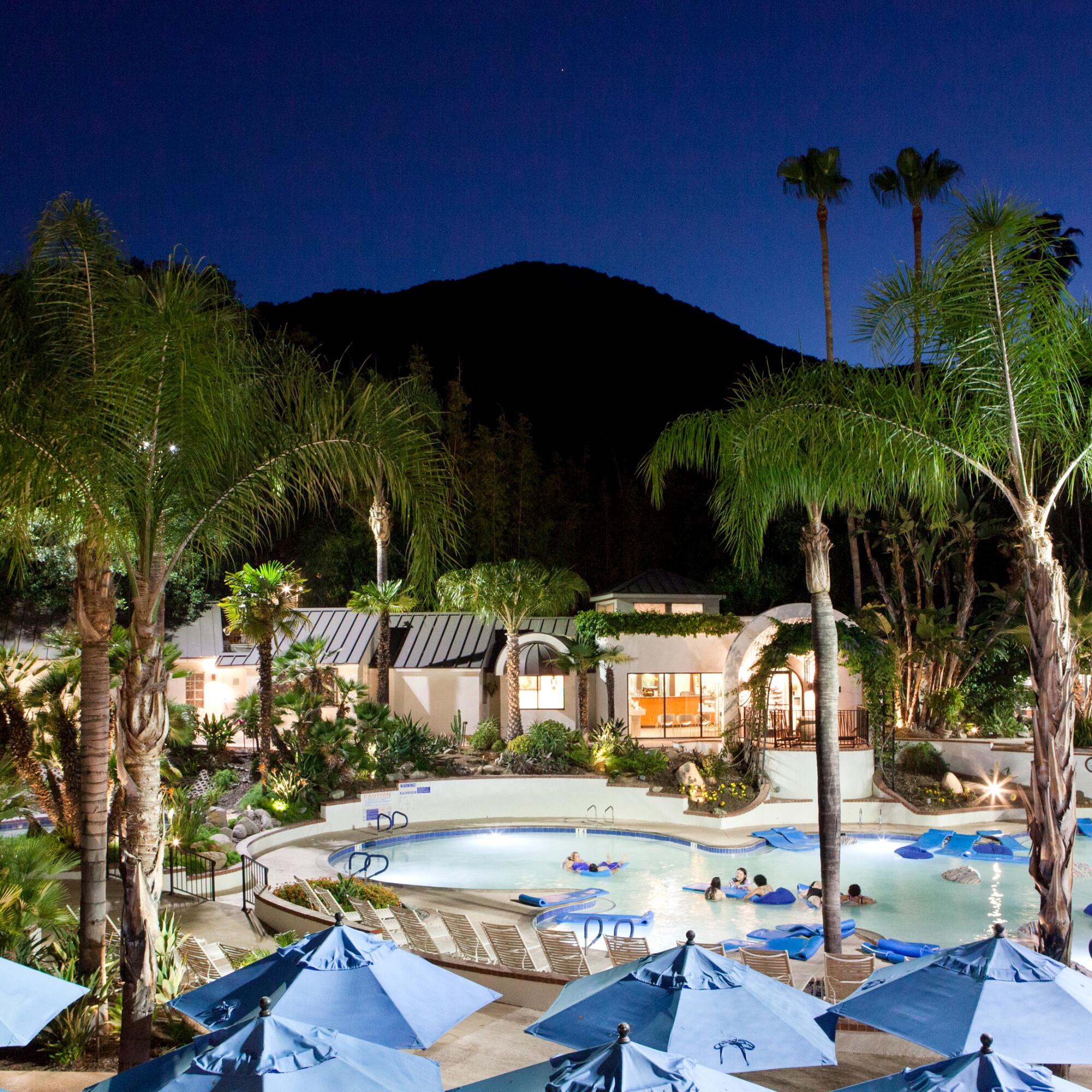 A pool at nighttime, surrounded by cabanas and palm trees.