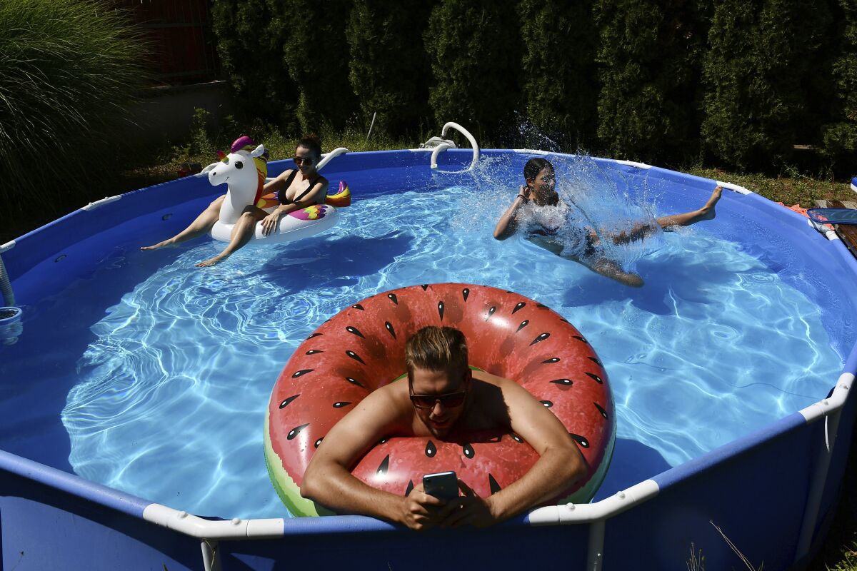 Barnabas types on his phone as he works from the home office as a pool in the garden while his wife, Rebeka, left and sister-in-law, Emma, cool down in Budapest, Hungary, Thursday, Aug. 4, 2022. Hungary has experienced several heat waves since mid June with day-time temperatures rising to 40 degrees Celsius and remaining at tropical levels through the night. In fact, the country recently shattered record for its hottest night ever when temperature of 25.4 C was measured in Budapest on July 26. (AP Photo/Anna Szilagyi)