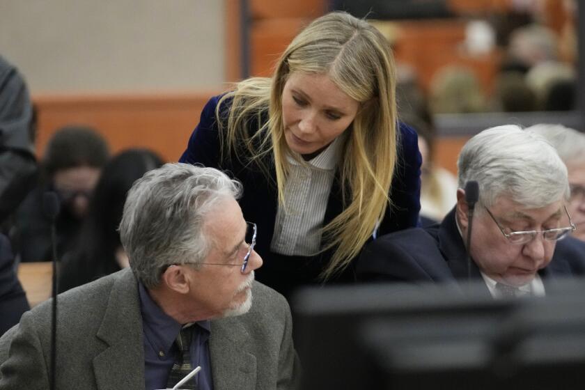 Gwyneth Paltrow wearing blue suit with white shirt,  leaning over to speak with seated man in gray suit