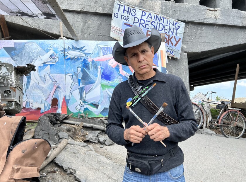 A man in a hat and bandolier with brushes holds two paintbrushes forming an X in front of a painting near a damaged bridge