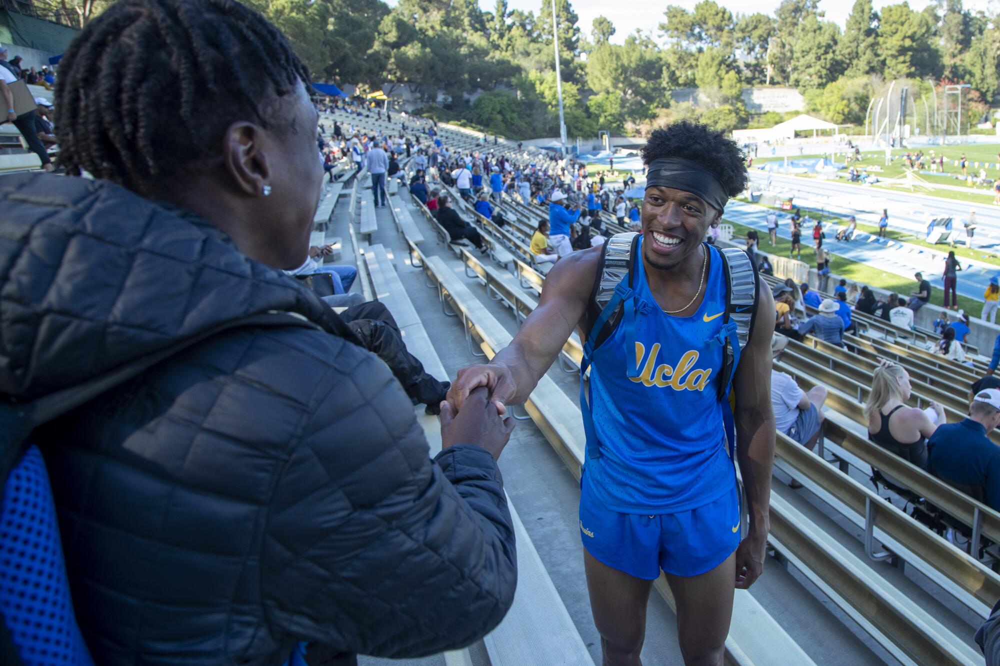 An athlete in UCLA runner's gear shakes hands in the stands with a friend 