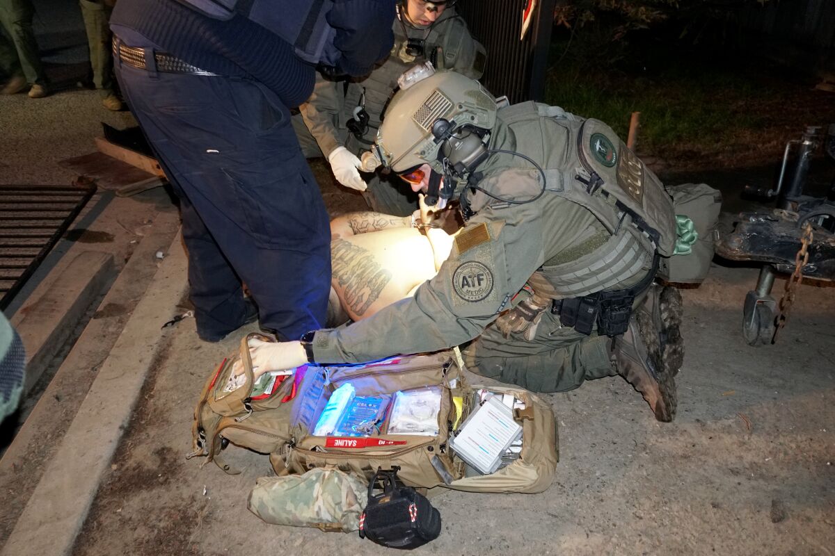 A man in tactical gear leans over a first aid kit next to a man lying on the ground.
