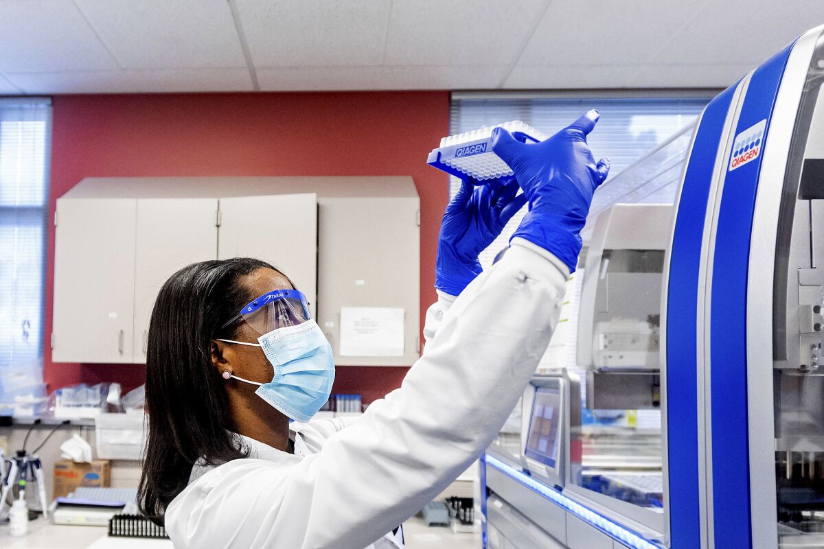 A woman a face shield, mask and gloves holds a rack of vials