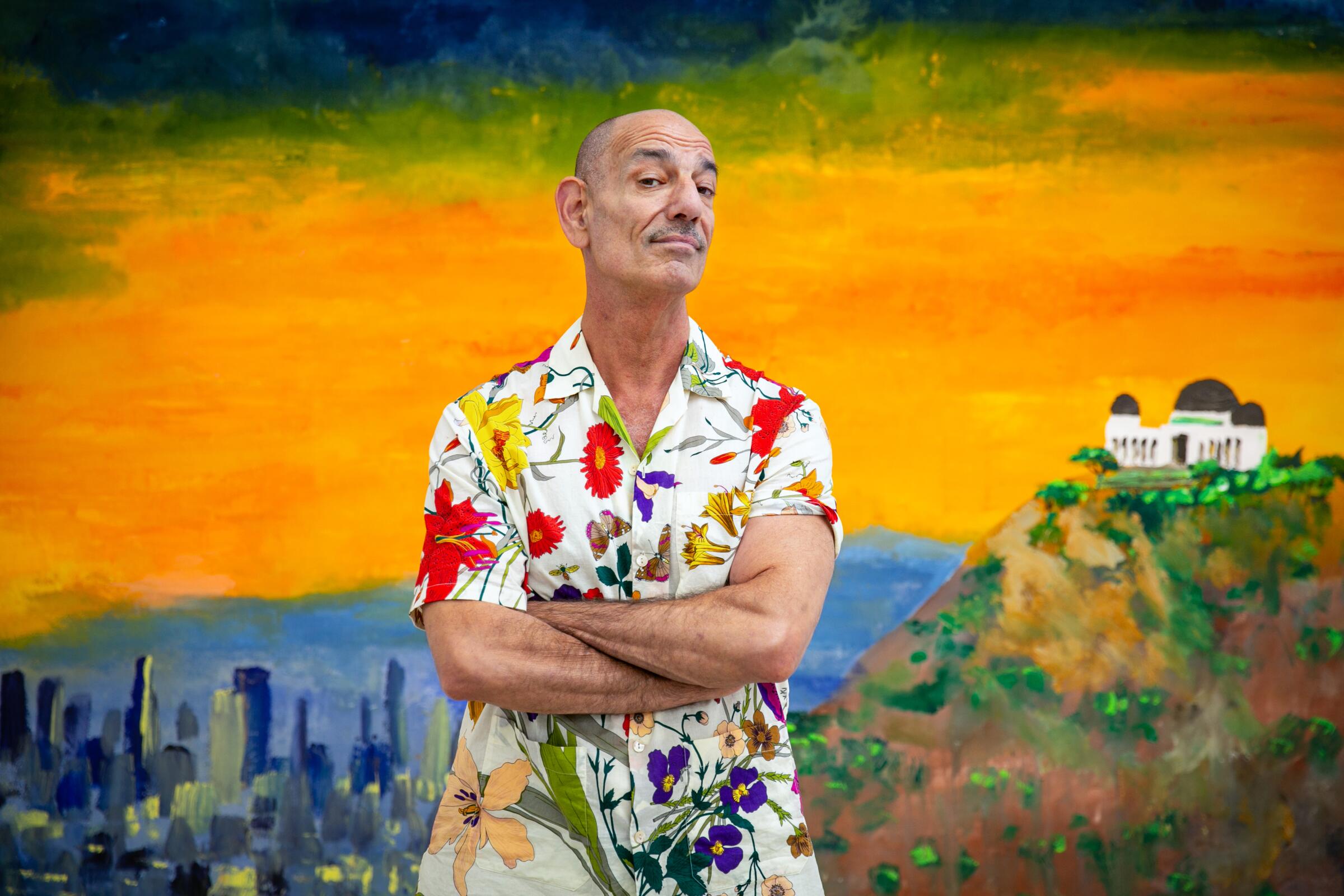 A man wearing a colorful floral shirt stands in front of a painted canvas of L.A. that features Griffith Observatory