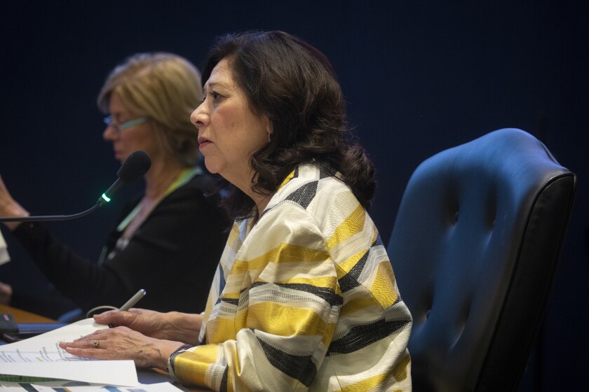 Supervisor Hilda Solis speaking into a microphone while holding paperwork and seated in an office chair